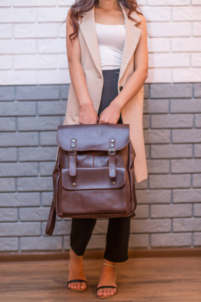 Женщина держит коричневый кожаный рюкзак в руке на фоне кирпичной стены. Мешок Unisex - Фото, изображение