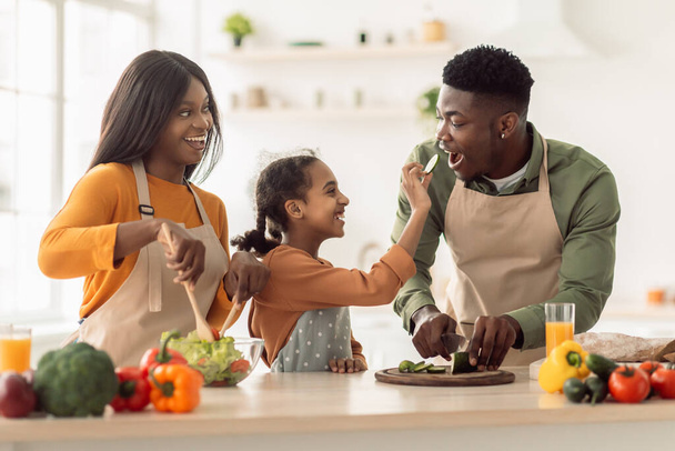 Black Family Cooking, Daughter Feeding Father While Making Salad Indoor - Photo, Image