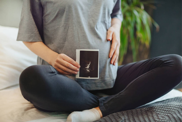 Cropped shot of pregnant woman holding pregnancy ultrasound while sitting in lotus pose on bed at home, expectant mother with baby sonogram. Pregnancy, maternity and antenatal care - Фото, изображение