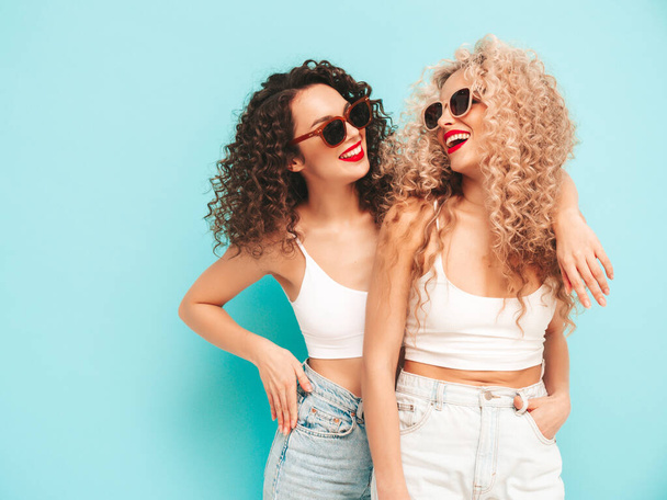 Two beautiful sexy smiling hipster women in red summer swimwear bathing suits. Trendy models with afro curls hairstyle having fun in studio. Hot female isolated on blue. In sunglasses - Fotoğraf, Görsel