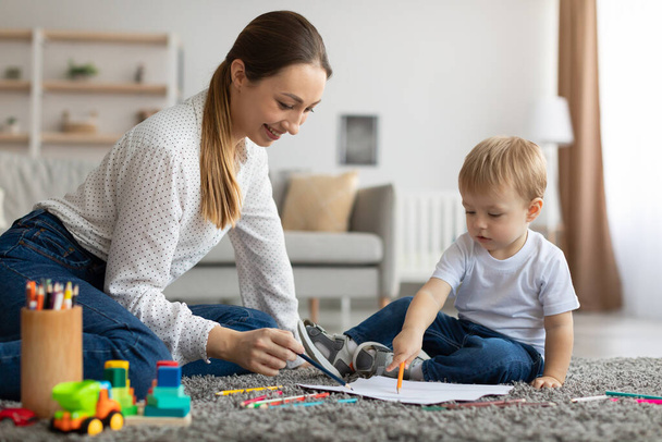 Vidám Y-generációs anya és kisgyermek fia ül a padlón, színes ceruzákkal és papírlapokkal rajzolva. - Fotó, kép