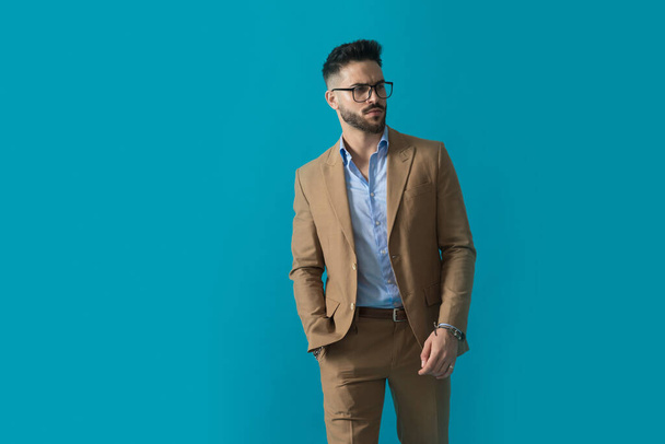 handsome businessman with glasses holding hands in pockets and looking to side while walking on blue background in studio - Fotoğraf, Görsel