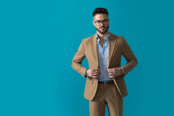 zelfverzekerde zakenman met bril knopen pak jas en op zoek naar kant op blauwe achtergrond in de studio - Foto, afbeelding