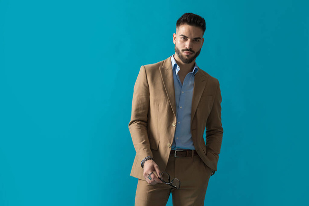 handsome young guy in beige suit holding hand in pocket, holding eyeglasses and posing in a cool manner on blue background in studio - Photo, Image