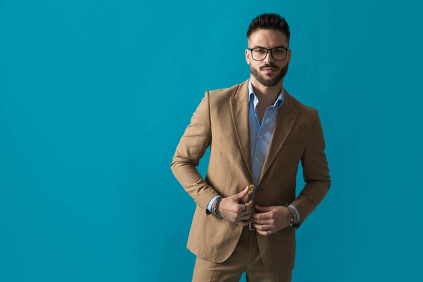attractive businessman with glasses buttoning suit and posing on blue background in studio - Photo, Image
