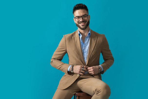 siedzący szczęśliwy młody człowiek w okularach uśmiecha się i dopasowuje garnitur siedząc na niebieskim tle w studio - Zdjęcie, obraz