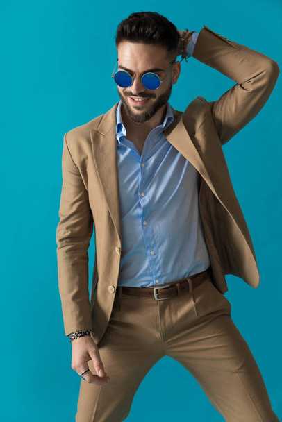 счастливый бизнесмен с бородой в ретро-очках и держа руку за голову, позируя в студии на синем фоне - Фото, изображение