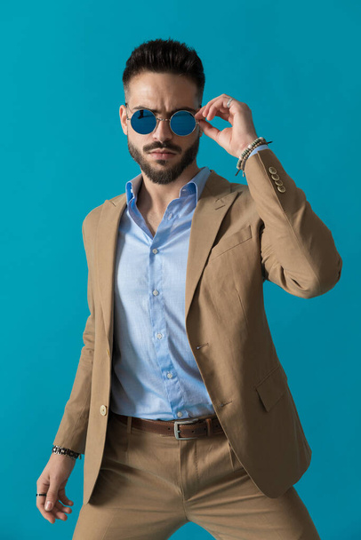 δροσερό νεαρός άνδρας της μόδας σε μπεζ κοστούμι στέκεται με έναν τρόπο μόδας, ενώ επιδιόρθωση γυαλιά μπροστά από το μπλε φόντο στο στούντιο - Φωτογραφία, εικόνα