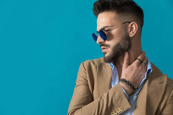 side view of sexy young mean with beard wearing retro sunglasses holding hand behind neck and looking to side on blue background in studio - Foto, immagini