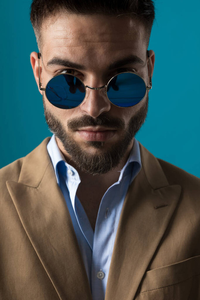 retrato de hombre barbudo atractivo con gafas de sol retro posando y mirando sobre gafas en fondo azul en el estudio - Foto, Imagen
