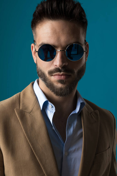 cool sexy biznesmen w beżowy garnitur noszenie okulary przeciwsłoneczne retro i pozowanie przed niebieskim tle w studio - Zdjęcie, obraz