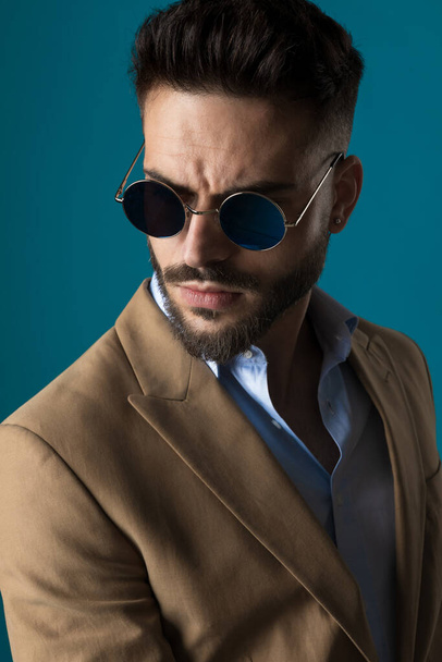 portrait of cool businessman wearing retro sunglasses and looking away while posing in a confident manner on blue background in studio - Foto, Imagem