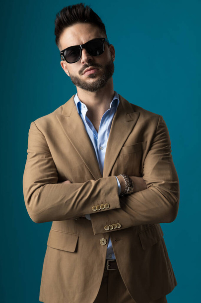 sebevědomý mladý podnikatel přes rameno a pózování v béžovém obleku se slunečními brýlemi na modrém pozadí ve studiu - Fotografie, Obrázek