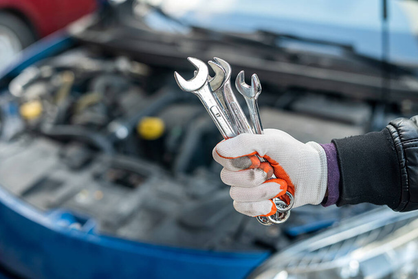 hand monteur in een speciale handschoen houdt een set van gereedschappen voor de auto in de buurt van de auto met een open motorkap. Autoreparatieconcept - Foto, afbeelding