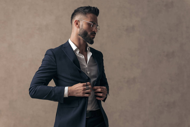 young handsome businessman with tough attitude is arranging his jacket, wearing eyeglasses and looking to side - Photo, Image