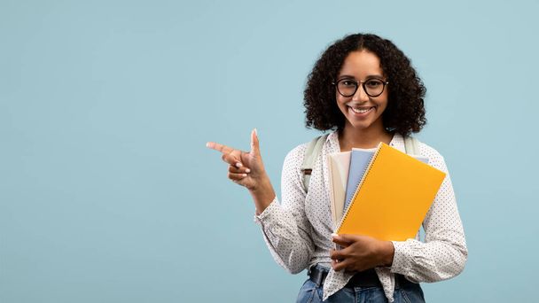 Allegra studentessa nera con zaino e materiali di studio che puntano da parte lo spazio per il tuo annuncio su sfondo blu - Foto, immagini