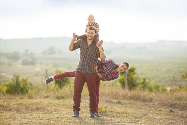 a young woman and a man with a little boy on their shoulders are standing in a vineyard . They are wearing plaid shirts - Fotoğraf, Görsel