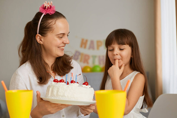 Beautiful young adult woman holding holiday dessert for her daughter birthday, little kid tasting cake, licking her finger, enjoying sugary delicious sweets. - 写真・画像