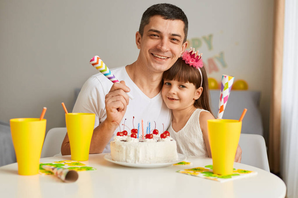 Portrait of attractive man, young father posing at home with his daughter, celebrating birthday with cake and candles, looking at camera with happy smile, expressing happiness. - Foto, Imagem