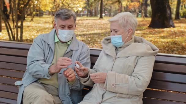 Starší pár v lékařských maskách v podzimním parku manželé dezinfikovat ruce s antiseptiky sedí na lavičce důchodci péče o zdraví osobní hygiena starý muž objetí manželka epidemie covid19 prevence koncept - Záběry, video