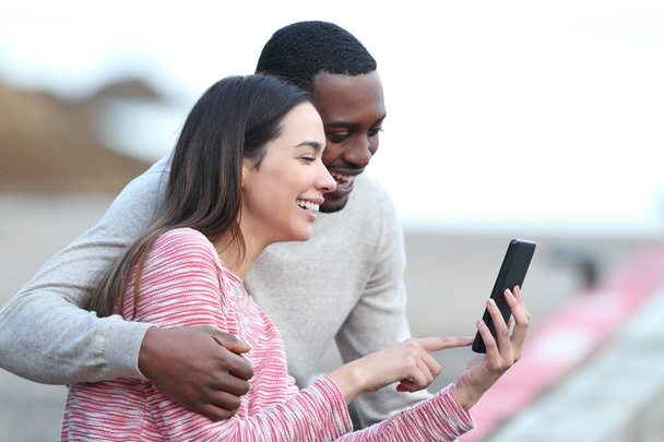 Happy interracial couple checking smart phone sitting outdoors - Фото, изображение