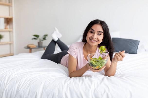 Hermosa mujer india joven comiendo ensalada de verduras mientras está acostado en la cama en casa, espacio libre. Dieta equilibrada de alimentos crudos - Foto, Imagen