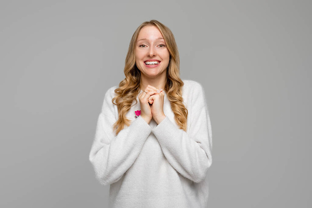 Touched, enthusiastic cute blonde woman gasping surprised and flattered, holding hands under chin, gazing camera thankful, stand pleased, wears white sweater, gray background - Photo, Image