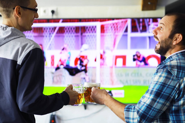 Hands horizontal holding lager beer glass and clinking on background of football game. Sport fans cheer up. Friends leisure lifestyle concept. - Photo, Image