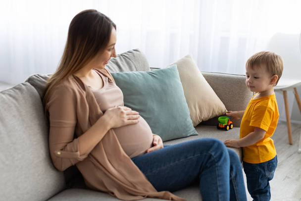 Щаслива вагітна мати проводить час зі своїм маленьким сином, сидячи на дивані і торкаючись її живота
 - Фото, зображення