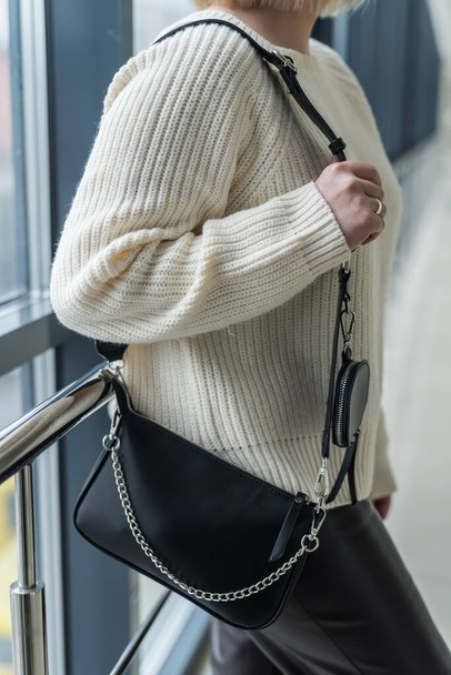 Trendy jonge meid in een warme gezellige trui pronkt met een stijlvolle zwarte leren tas. Het concept van stijlvolle accessoires - Foto, afbeelding