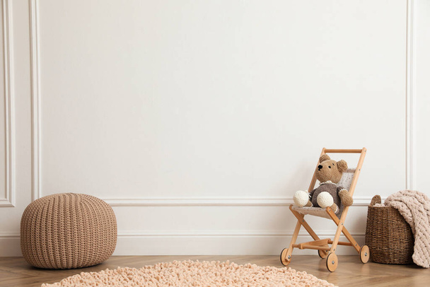 Toy stroller with bear, pouf and wicker basket near white wall in child room. Interior design - Φωτογραφία, εικόνα