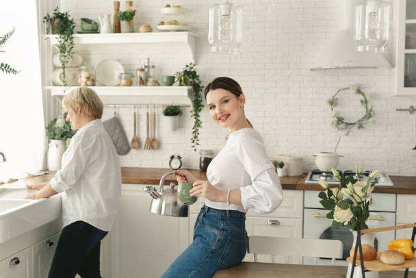Adult daughter pours tea from a teapot into a mug while her elderly mother washes dishes in the kitchen sink - Foto, afbeelding