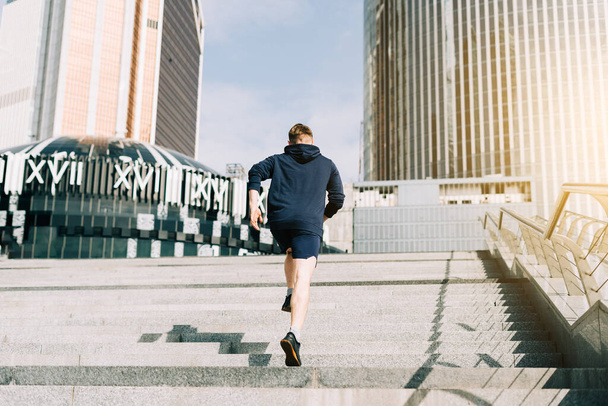 Молодой спортсмен бегает вверх и вниз по городской лестнице летом на утренней пробежке, на фоне городской улицы. Спортивная подготовка. Тренажерный зал на свежем воздухе, прогулка на свежем воздухе - Фото, изображение