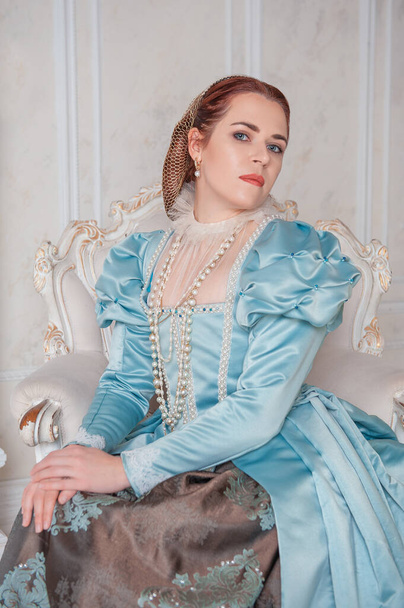 Mooie jonge vrouw in middeleeuwse stijl blauwe jurk zittend in de armchai - Foto, afbeelding