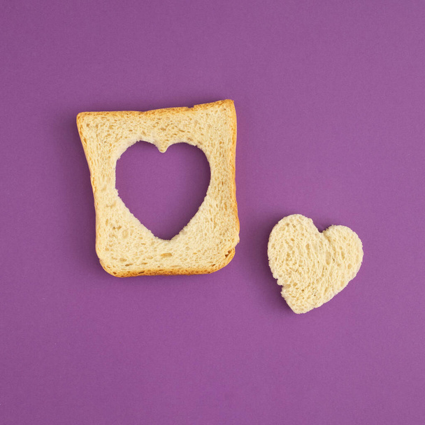 Composition with toasted bread with a slice in the shape of a heart against a purple background. Minimal love or food concept. Flat lay. - Fotoğraf, Görsel