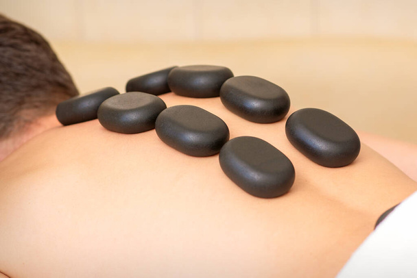 Massage mit heißen Steinen. Kaukasischer junger Mann bekommt im Wellness-Salon eine Hot-Stone-Massage auf dem Rücken - Foto, Bild
