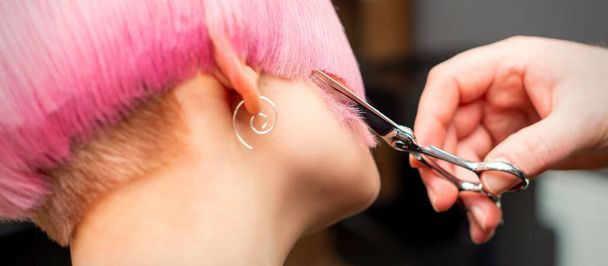 Vrouwelijk haar knippen. Hand van een kapper knippen korte roze haar van jonge blanke vrouw in de kapsalon - Foto, afbeelding