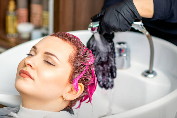 Washing dyed female hair. Professional hairdresser washes pink color paint off of a customer. Young caucasian woman having her hair washed in a beauty salon - Photo, Image