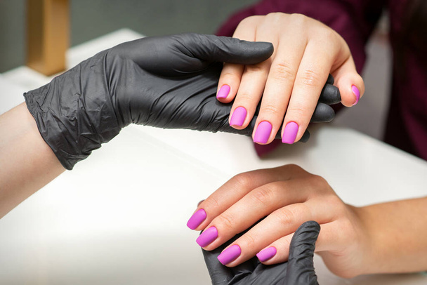 Examen de las uñas arregladas. Las manos del maestro de la manicura en guantes negros examinando las uñas rosas femeninas en el salón de manicura - Foto, imagen