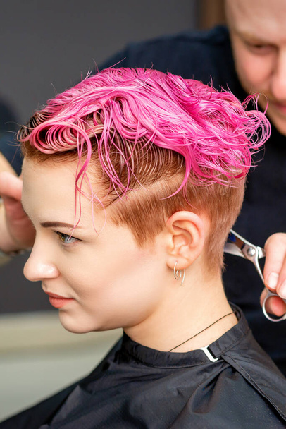 Femme ayant une nouvelle coupe de cheveux. Un coiffeur masculin coupe les cheveux courts teints en rose avec des ciseaux dans un salon de coiffure - Photo, image