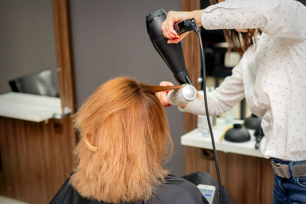 Ξερά μαλλιά στο κομμωτήριο. Γυναίκα κομμώτρια στυλίστας στεγνώνει τα μαλλιά με στεγνωτήρα μαλλιών και στρογγυλό πινέλο κόκκινα μαλλιά μιας γυναίκας σε ένα σαλόνι ομορφιάς - Φωτογραφία, εικόνα