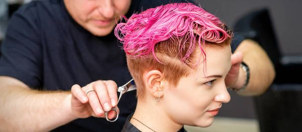 Egy nőnek új frizurája van. A férfi fodrász vágja festett rózsaszín rövid haj ollóval egy fodrászszalonban - Fotó, kép