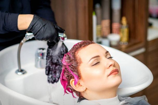 Washing dyed female hair. Professional hairdresser washes pink color paint off of a customer. Young caucasian woman having her hair washed in a beauty salon - Photo, Image