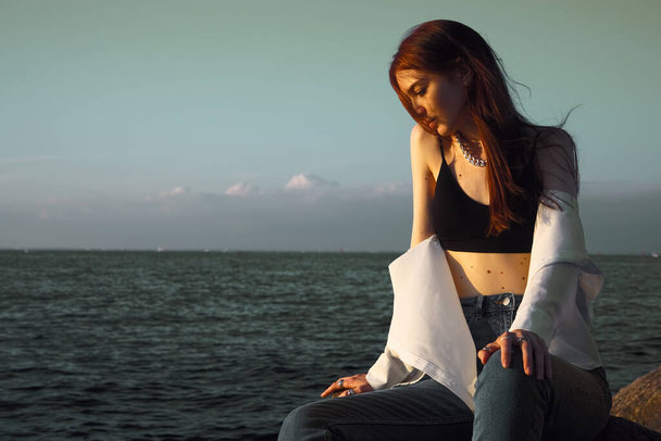Portrait of young dreaming woman on stones near sea. relaxing girl is resting on coast, enjoying outdoor recreation. - Φωτογραφία, εικόνα