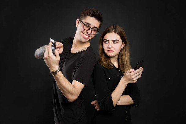Portrait of a young couple standing with mobile phone, man is playing games on mobile phone while angry girl standing near and looking at camera isolated over black background. - Foto, Bild