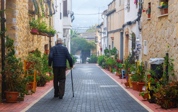 Achteraanzicht van een oudere man die met een wandelstok langs een prachtige straat met planten loopt, in Llber (Alicante, Spanje)) - Foto, afbeelding