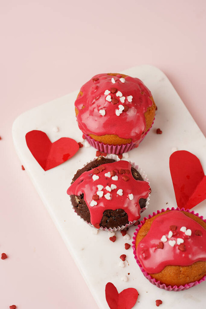 Três muffins de chocolate e branco caseiros com cobertura vermelha e decoração de açúcar vermelho e branco compartilhada pelo coração na placa de mármore na superfície rosa - Foto, Imagem