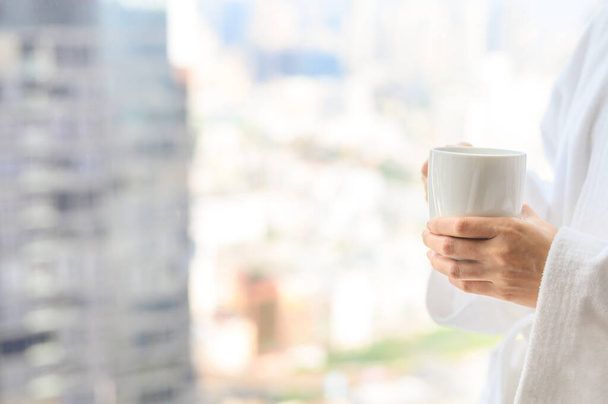 Vista lateral de la mujer en albornoz de pie cerca de la ventana mientras sostiene la taza de café blanco y disfrutar de una maravillosa mañana en el hotel, separándose del trabajo - Foto, Imagen