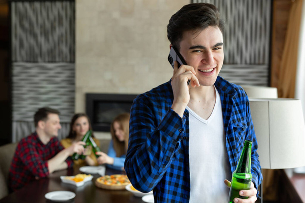 Young man drinks beer from a bottle, talks on the phone and looks out the window of a cafe against the background of his friends who are sitting at the table and eating pizza - Foto, Imagen