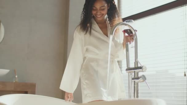 仕事がリラックスした後。若いアフリカ系アメリカ人の女性がワイングラスを楽しみ、自宅で浴槽に水を取り、ショットを追跡 - 映像、動画